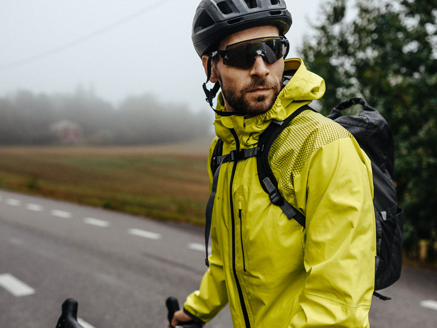 Keltaiseen takkiin ja kypärään pukeutunut mies seisoo polkupyörän kanssa tienvarressa ja katsoo sivulle sumuisena päivänä.
