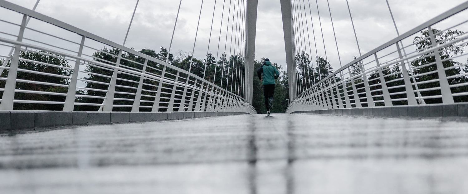 Vihreään takkiin pukeutunut henkilö juoksee yksin märällä, modernilla riippusillalla pilvisen taivaan alla.