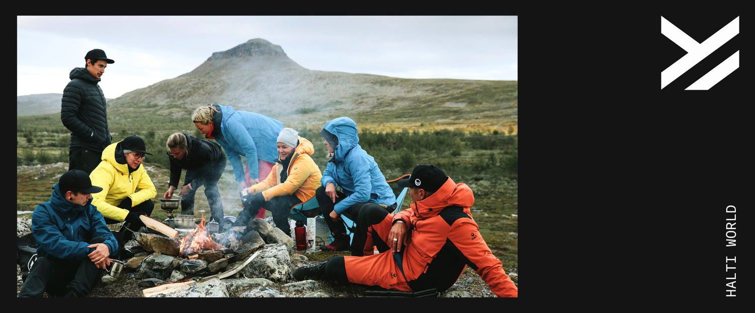 Ryhmä ulkoiluvaatteisiin pukeutuneita ihmisiä istuu ja seisoo nuotion ympärillä kallioisella maastolla, jonka taustalla on vuori.