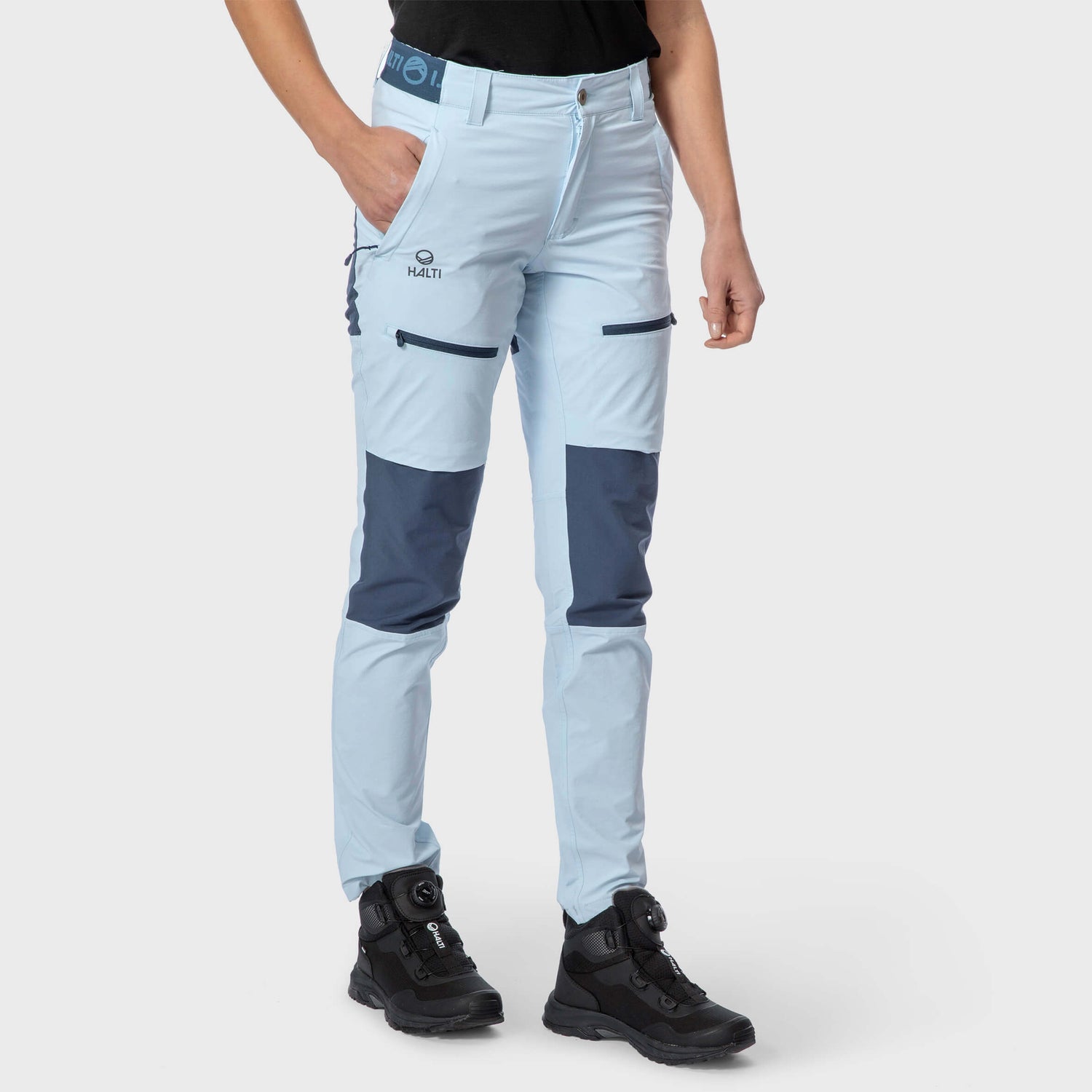 Henkilö, jolla on vaaleansiniset retkeilyhousut, joissa on tummansiniset polvilaastarit, ja mustat retkeilykengät, seisoo tavallista taustaa vasten.