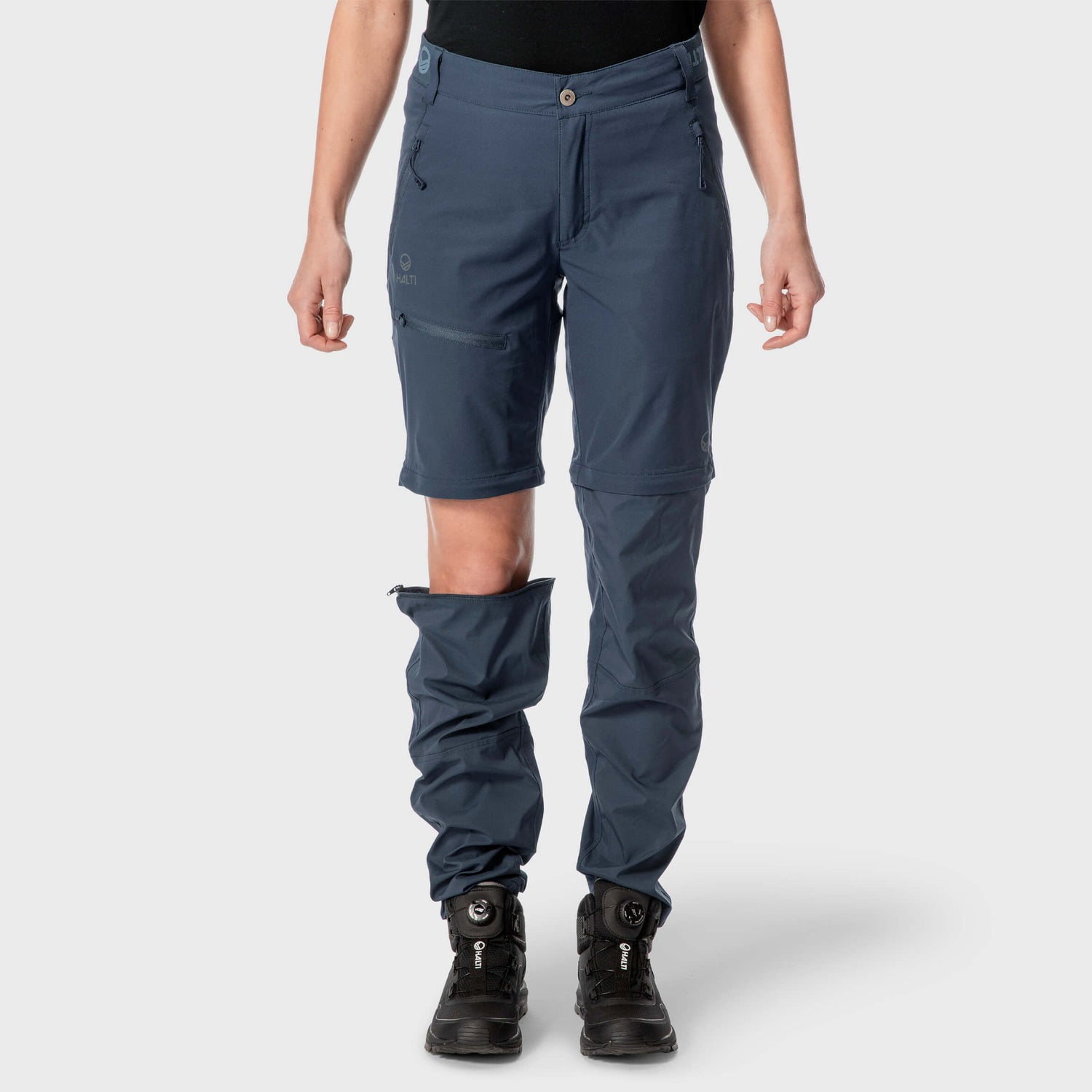 Henkilöllä on yllään siniset vetoketjulliset housut, joiden toinen jalka on muutettu shortseiksi, sekä mustat vaelluskengät, taustana tavallinen tausta.