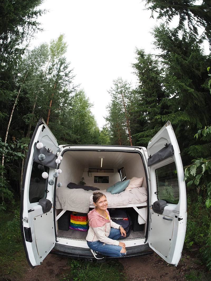 Henkilö istuu metsäiselle alueelle pysäköidyn valkoisen pakettiauton, jossa on sänky, avoimilla takaovilla.