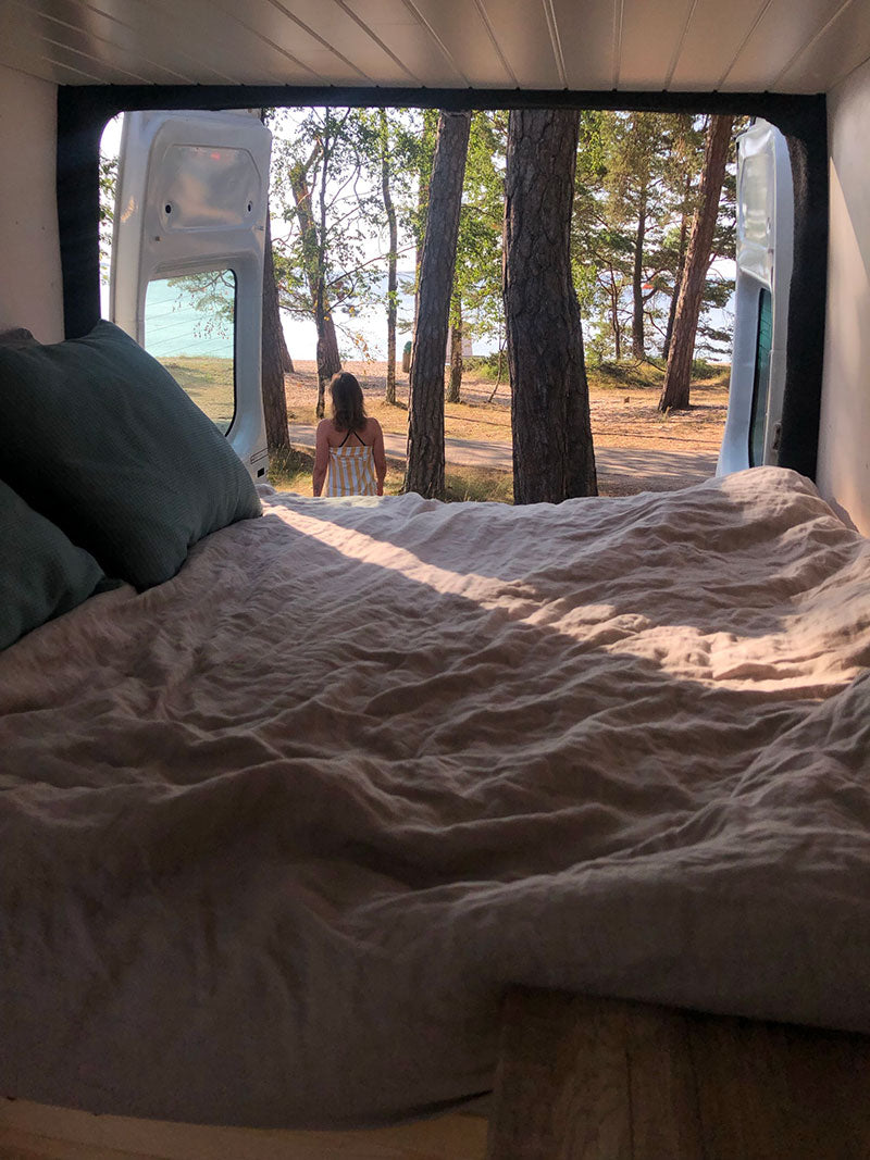 Näkymä pakettiauton sisältä, jossa on avoimet ovet, ja jossa henkilö istuu ulkona puiden ja taustalla näkyvän järven läheisyydessä.