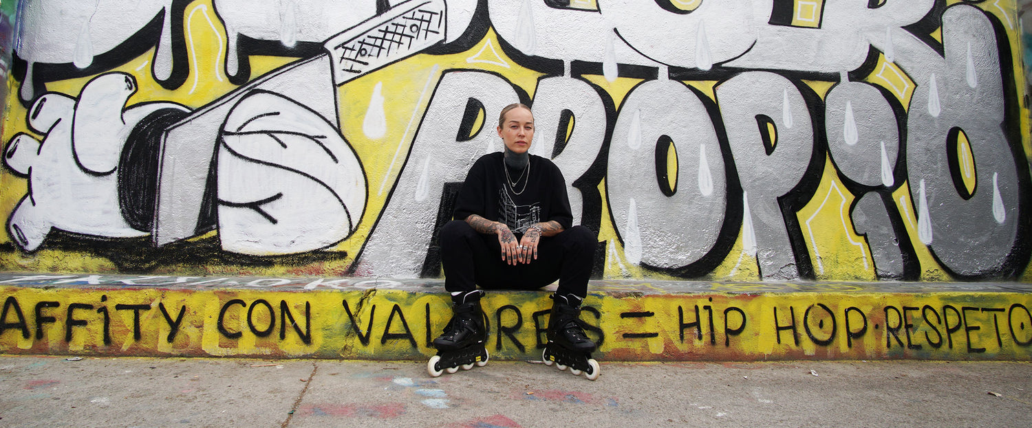 Mustiin vaatteisiin pukeutunut henkilö istuu reunalla värikkään graffitin peittämän seinän ja maan edessä.