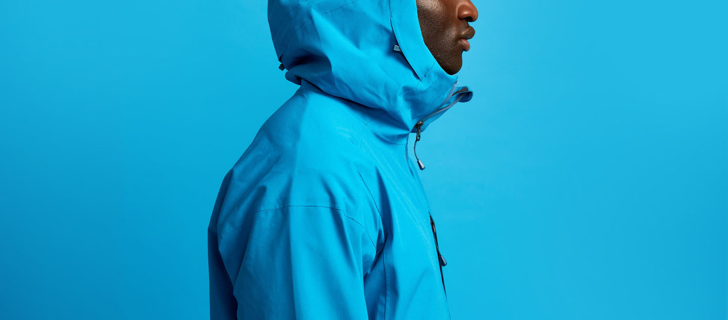 Siniseen hupulliseen takkiin pukeutunut henkilö seisoo profiilikuvassa yhtenäistä sinistä taustaa vasten.