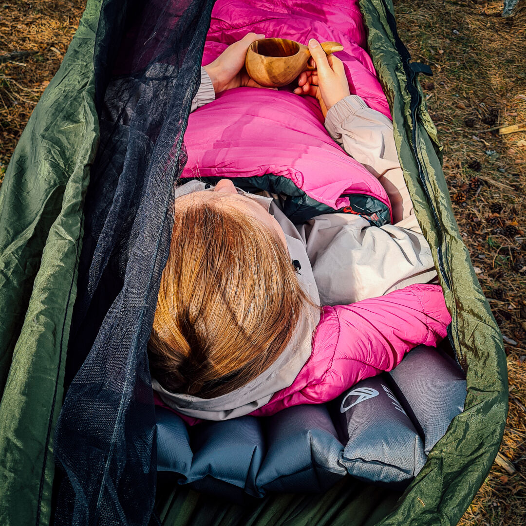 Vaaleanpunaiseen Halti Dreamcatcher 30 M makuupussiin pukeutunut henkilö makaa vihreässä riippumatossa ulkona puinen muki kädessään.