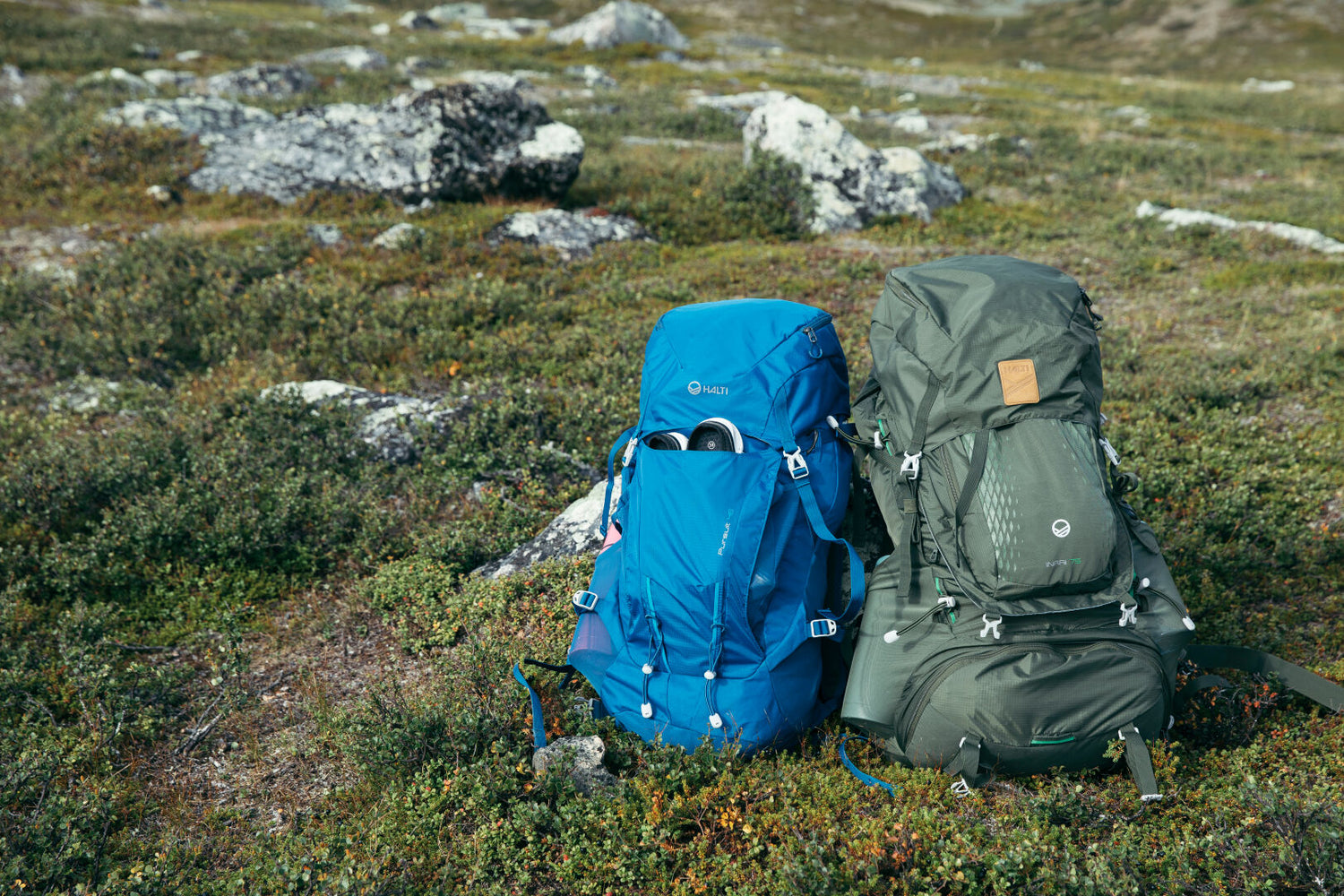 Kaksi Halti Inari 75 Rinkka -reppua, sinistä ja vihreää, kierrätetystä polyesteristä, on ruohikolla kivien takana.
