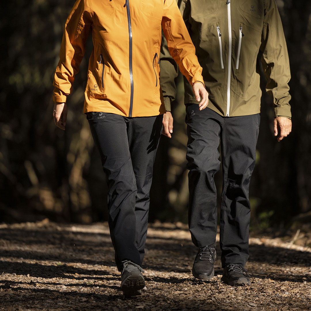 Kaksi ihmistä kävelee metsäpolulla Halti Leisti DX Kuorihousut Naisten alueella; vain heidän alavartalonsa näkyvät.