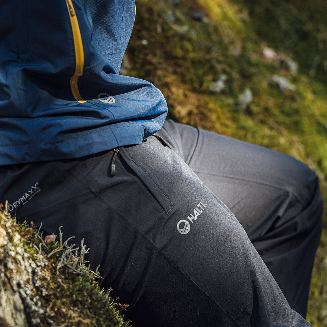 Mies, jolla on Halti Vuoksi DX Kuorihousut, istuu ulkona sammaloituneella maalla, DRYMAXX-logo näkyvissä.