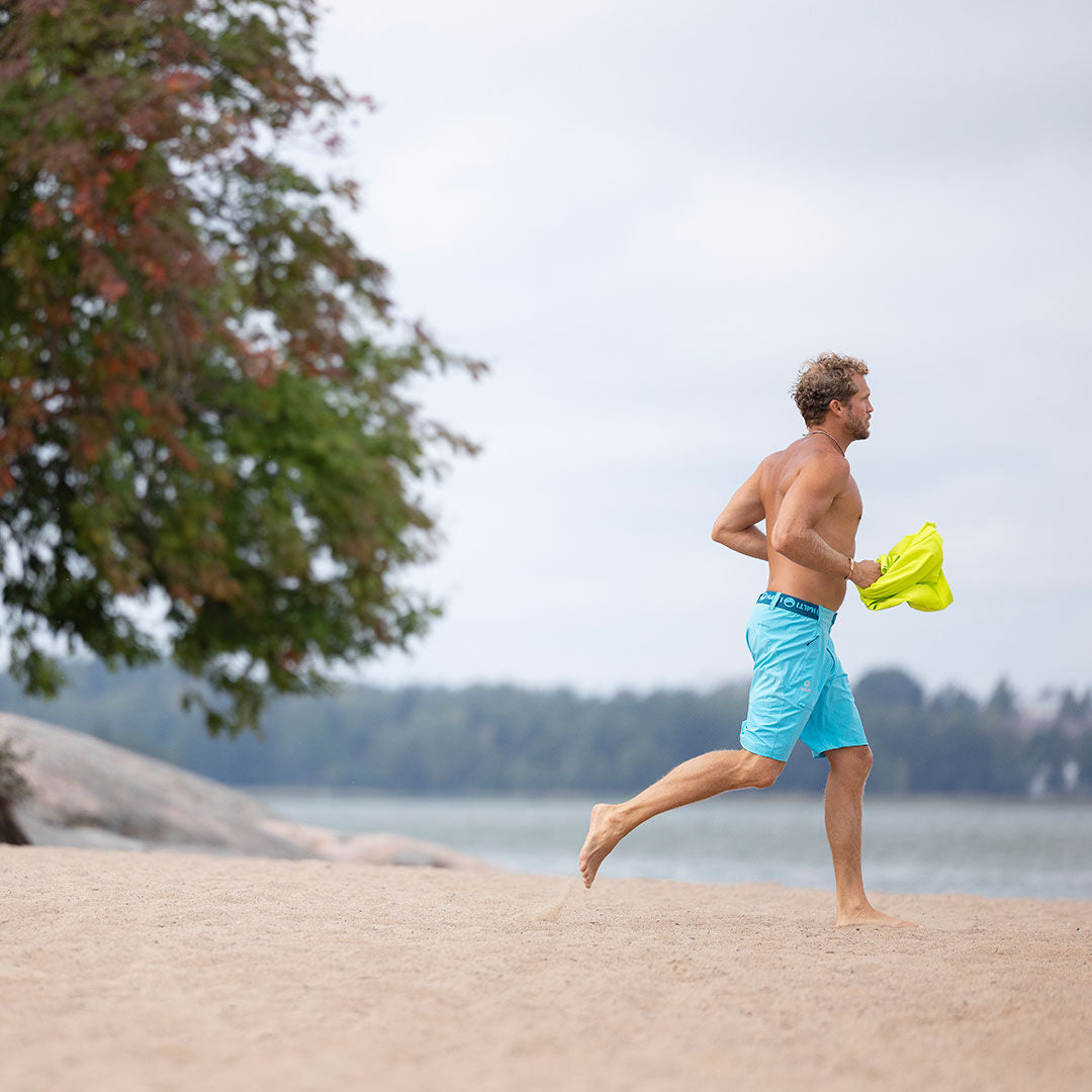 Mies juoksee paljain jaloin hiekkarannalla sinisissä Halti Pallas Miesten X-stretch Lite -shortseissa keltainen paita kädessään.