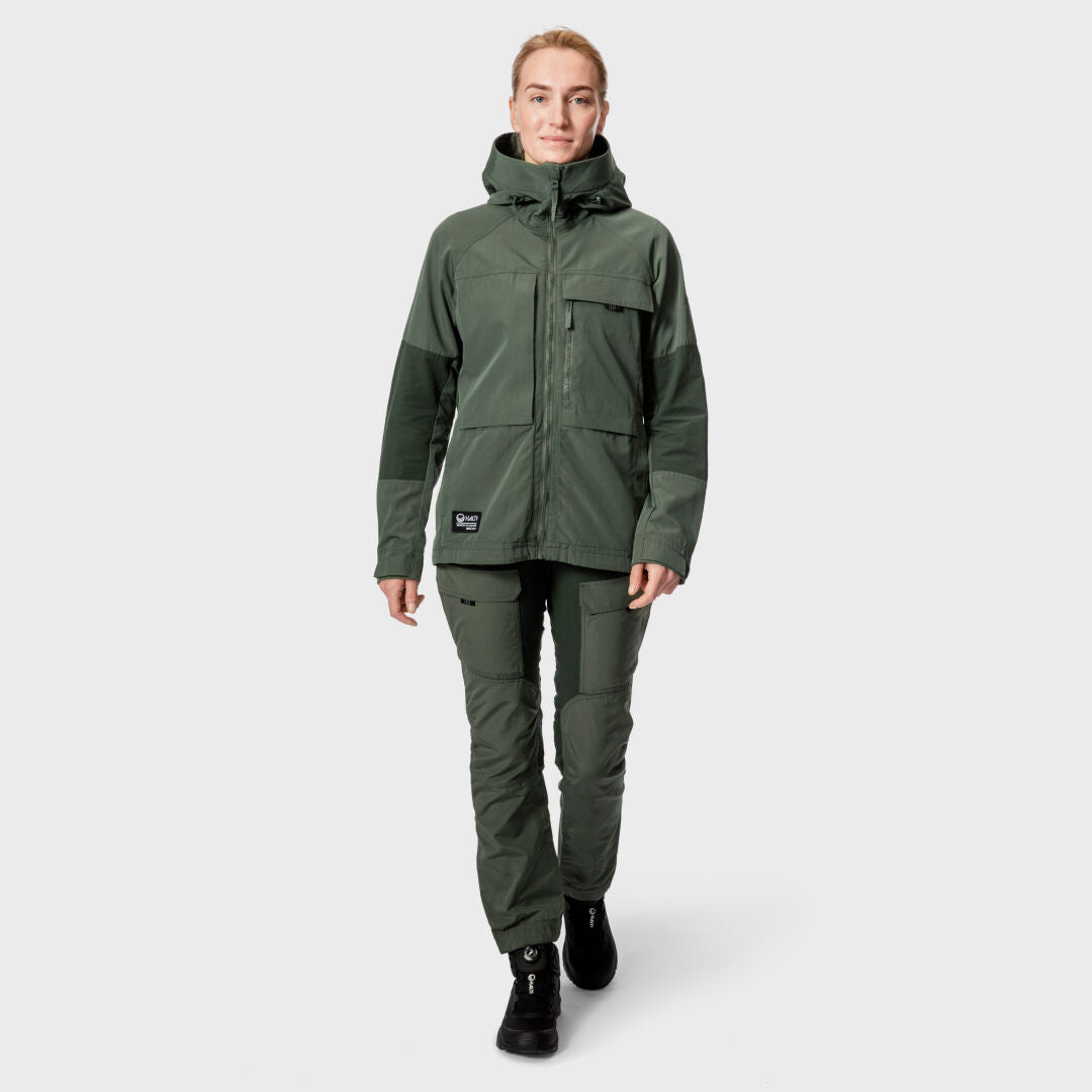 Henkilöllä on yllään Halti Hiker Naisten Ventilated Retkeilyhousut, vihreä takki ja mustat saappaat vaaleaa taustaa vasten.