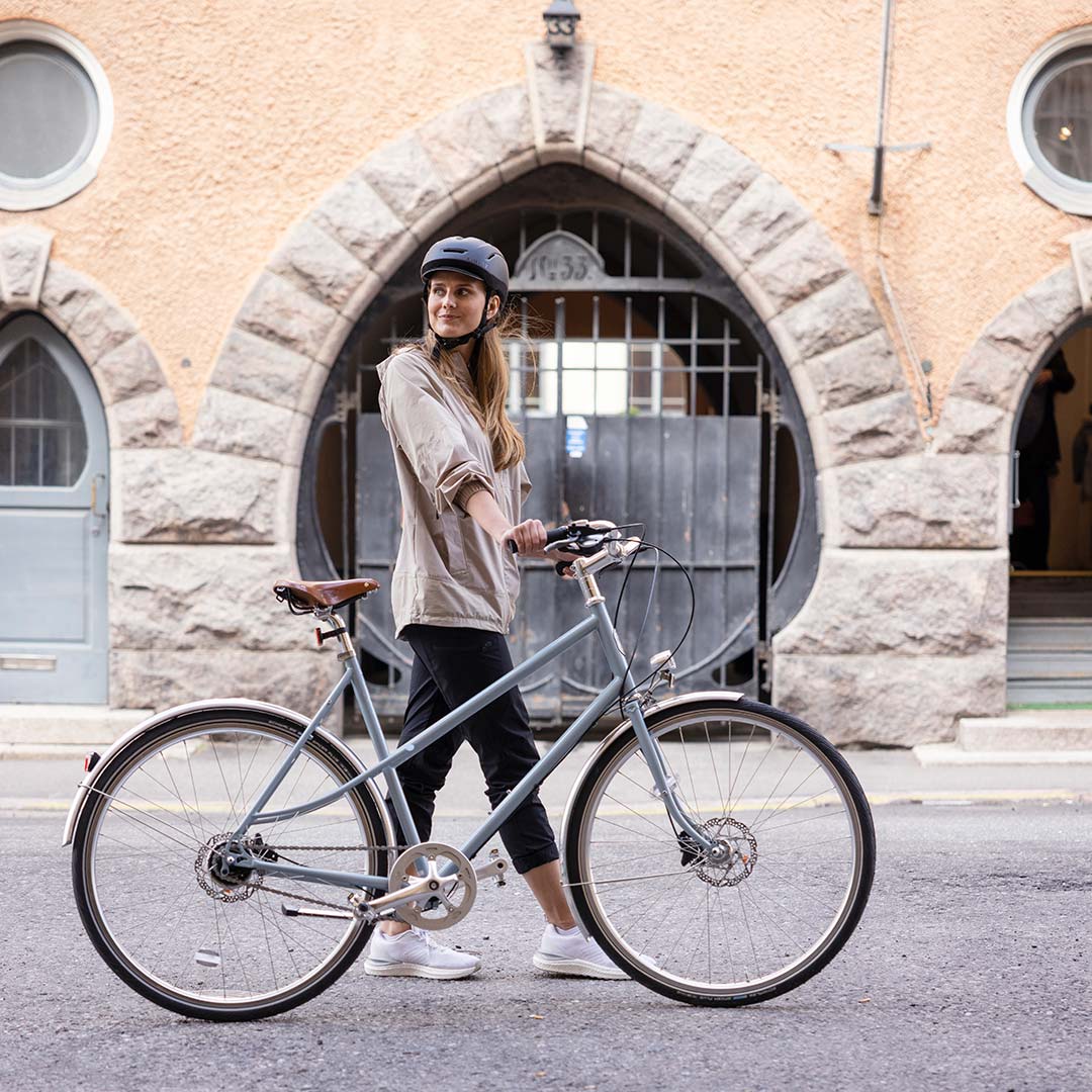 Halti Pallas Naisten Jogger Lite Takki -kengissä oleva nainen seisoo pyöränsä vieressä kypärä päässään kivitalon ulkopuolella.