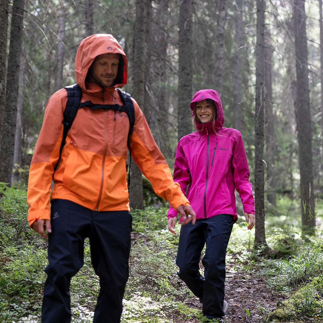 Kaksi ihmistä Halti Vuoksi DX Kuorihousut Miesten kävelee metsäpolulla - toinen pukeutuu oranssiin, toinen vaaleanpunaiseen - puiden seassa.