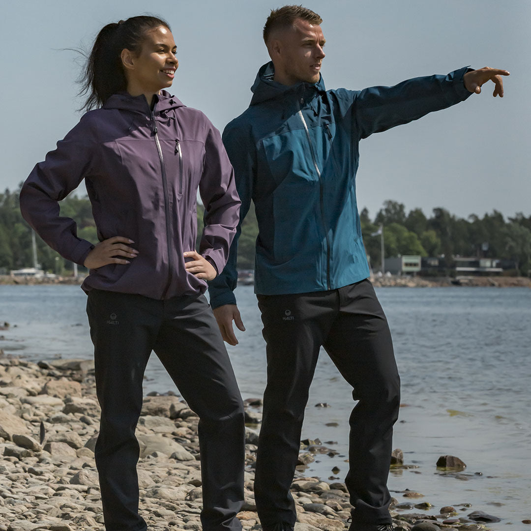 Kaksi ulkoilutakkiin ja Halti Leisti DX Kuorihousut Naisten vaatteisiin pukeutunutta ihmistä seisoo kivikkoisella järvenrannalla; mies osoittaa eteenpäin.