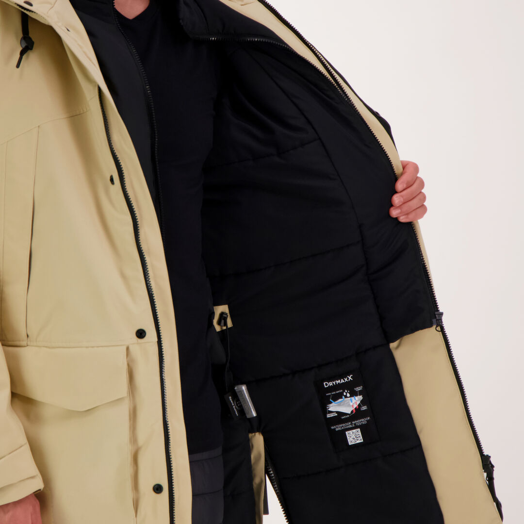 Beigeen takkiin pukeutunut henkilö pitää takkia auki, jotta sen sisältä näkyy Halti Bergga DrymaxX Talvitakki Miesten etiketti.