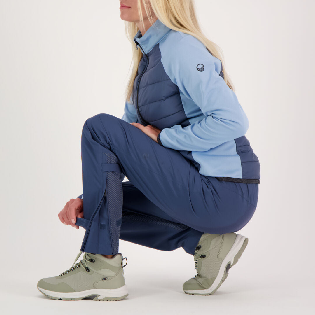 Pitkät vaaleatukkainen nainen kyykistyy sinisessä takissa, Halti Liike Naisten 3L DrymaxX Urheiluhousut ja kevyissä vaelluskengissä.