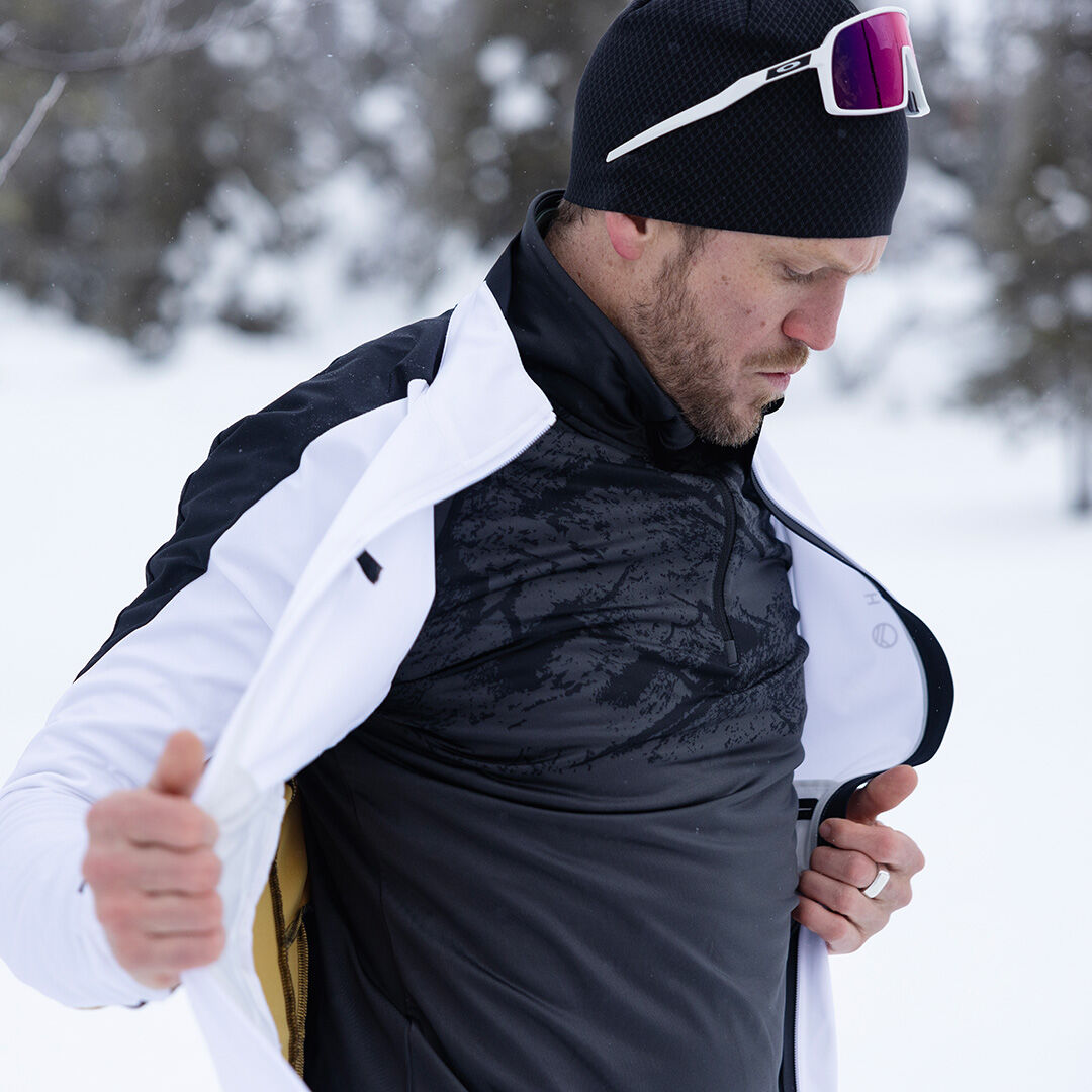 Talviurheiluvaatteissa oleva mies säätää Halti Crosser Paita Miesten -takkiaan ulkona aurinkolasit ja pipo päässä.