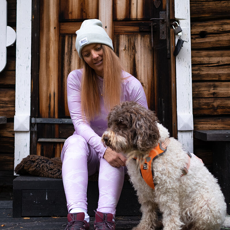 Pehmeällä harjatulla vuorella varustettua Halti Windfire Kerrasto Naisten -takkia käyttävä nainen istuu puisilla portailla oranssiin valjaisiin pukeutuneen koiran vieressä.