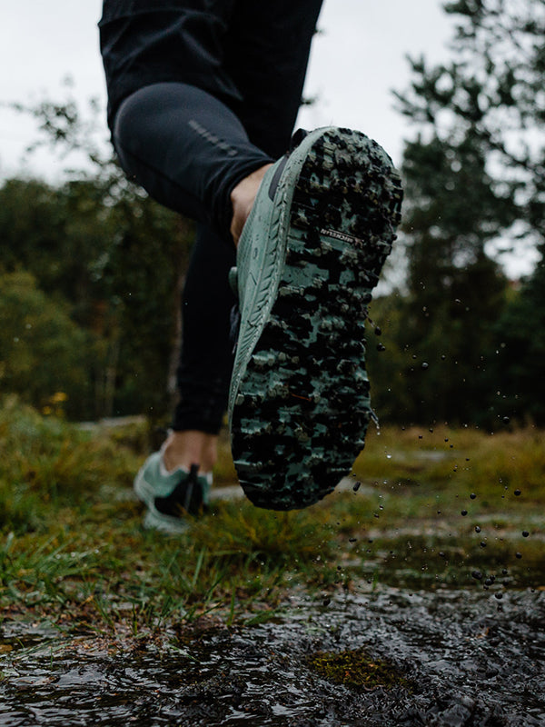 Henkilö, jolla on tummat urheiluhousut ja vihreät juoksukengät, astuu märällä, mutaisella ulkoilureitillä, jossa vesi roiskuu ja kengänpohjan kulutuspintakuvio näkyy. Taustalla näkyy puita.