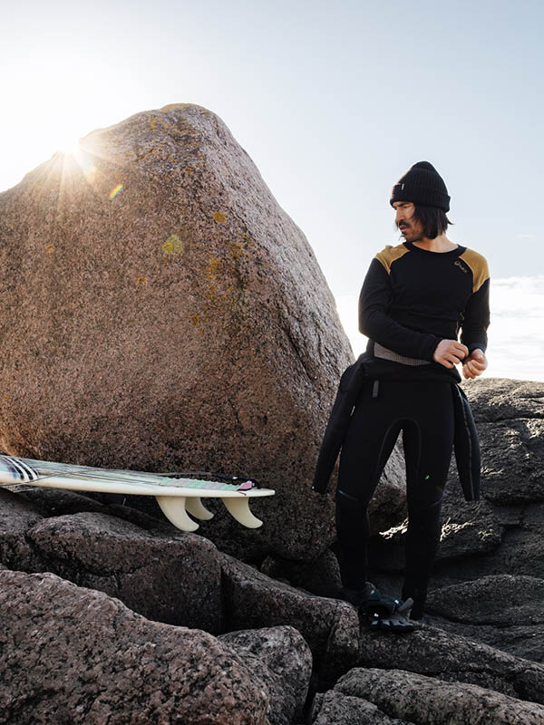Kylpypuku ja pipo päässään oleva henkilö seisoo kivikkoisella maastolla suuren lohkareen vieressä, ja surffilauta lepää kivillä hänen vieressään. Auringonvalo paistaa lohkareen takaa.