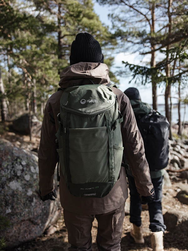 Kaksi ulkoiluvaatteisiin ja reppuihin pukeutunutta henkilöä kävelee metsäisellä alueella, jossa on kiviä ja puita. Edessä olevalla henkilöllä on vihreä Halti-reppu ja hänellä on musta pipo.