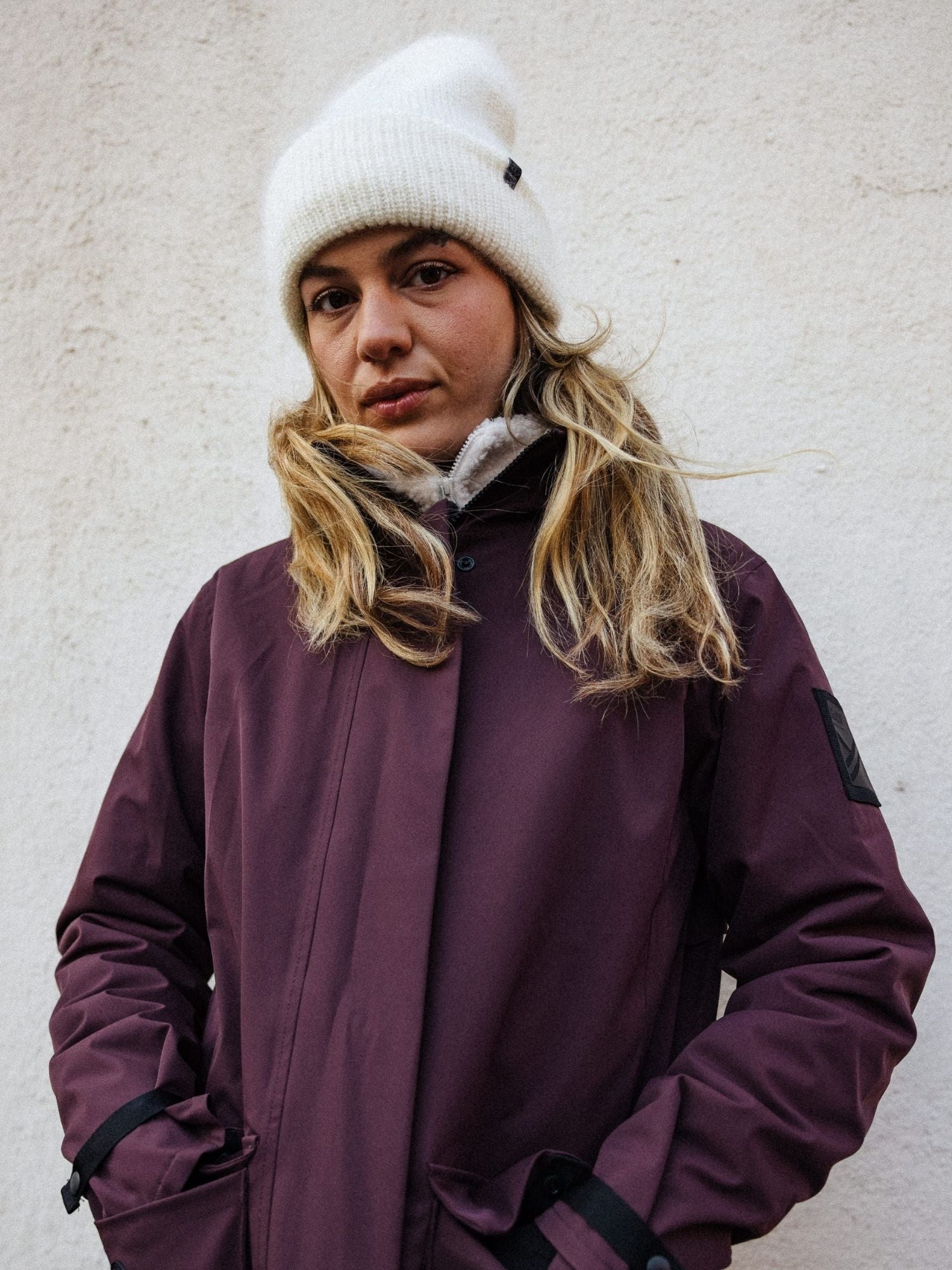 Pitkät vaaleat hiukset, valkoinen neuletakki ja ruskeankeltainen talvitakki, seisoo valkoista seinää vasten kädet takin taskuissa.