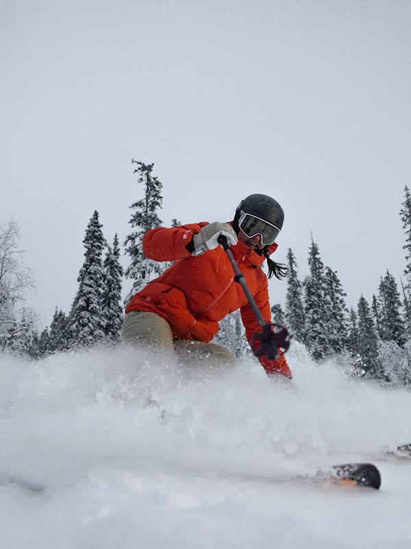 Henkilö, jolla on oranssi takki, beiget housut ja musta kypärä, hiihtää alamäkeen tuoreessa lumessa, taustalla ikivihreitä puita ja pilvinen taivas.