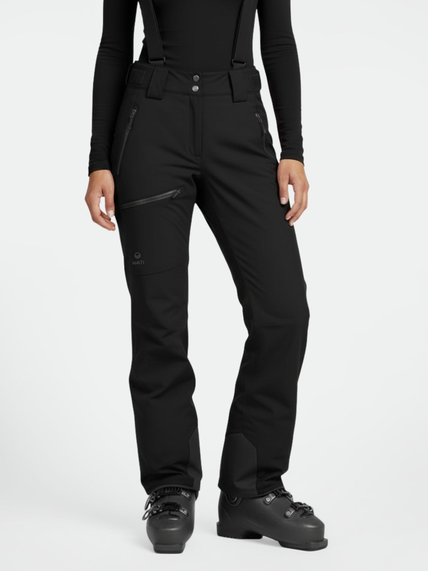 Henkilöllä on Halti Carvey DrymaxX Lasketteluhousut Naisten housut henkseleillä, musta toppi ja hiihtokengät valkoisella taustalla.