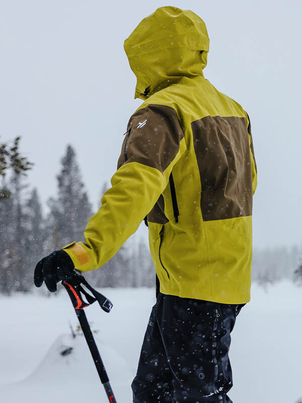 Henkilö, jolla on keltainen ja ruskea Halti Rizle 3L DX Laskettelutakki Unisex, seisoo lumessa hiihtosauvat kädessään ja katsoo poispäin.