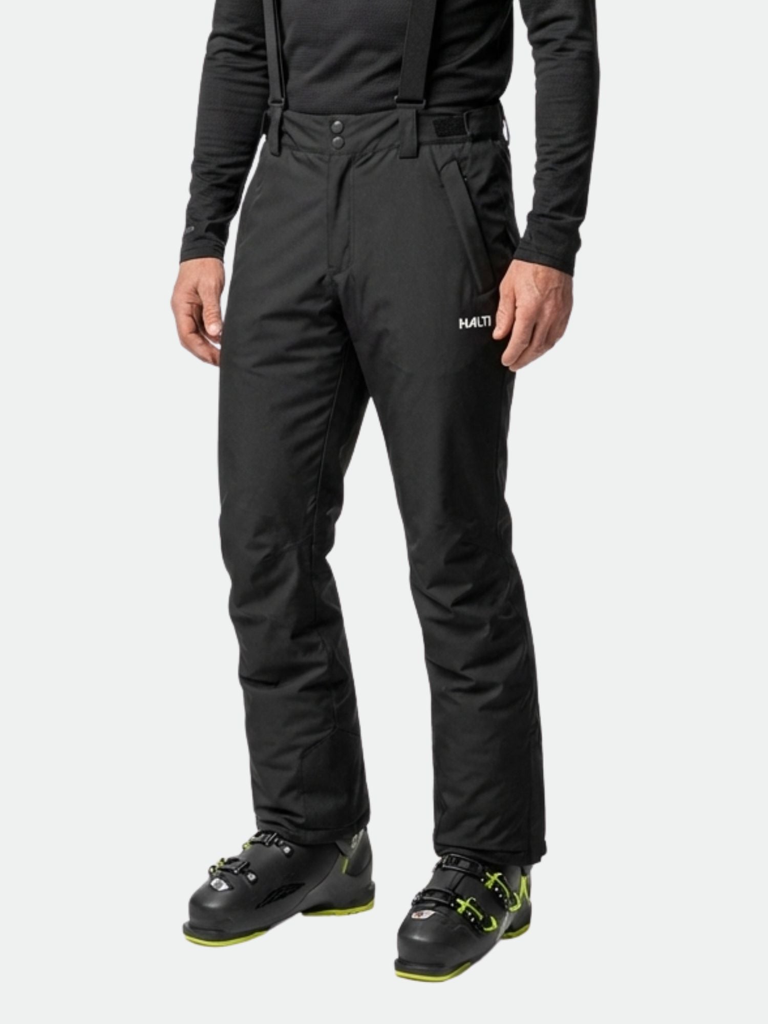 Henkilöllä on mustat Halti Glades II DX -hiihtohousut, joissa on henkselit, musta toppi ja vihreällä korostetut hiihtokengät.