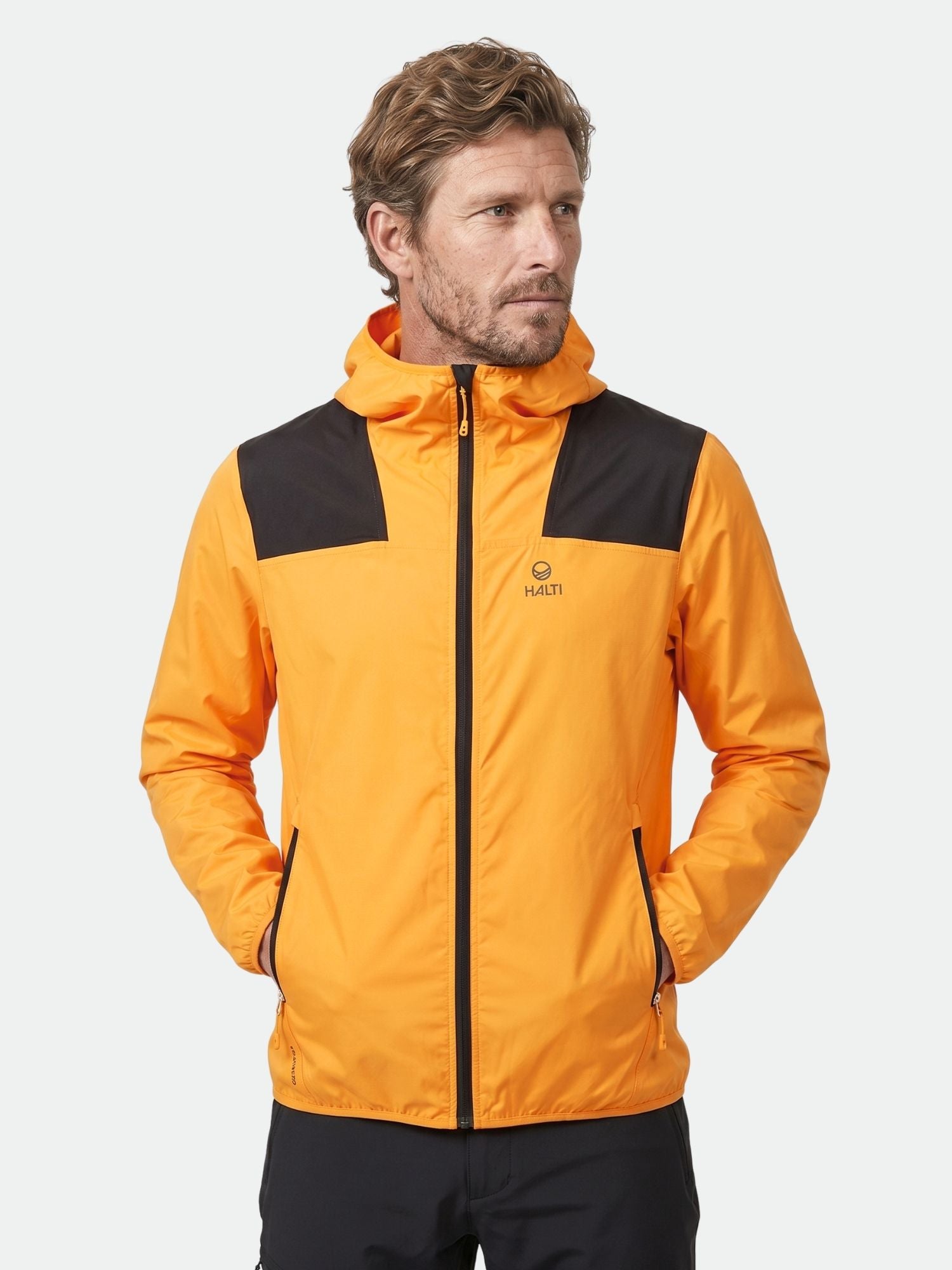 Miehellä Halti Pallas Miesten Lite X-stretch Ulkoilutakki, oranssi takki mustilla korostuksilla, kädet taskuissa.