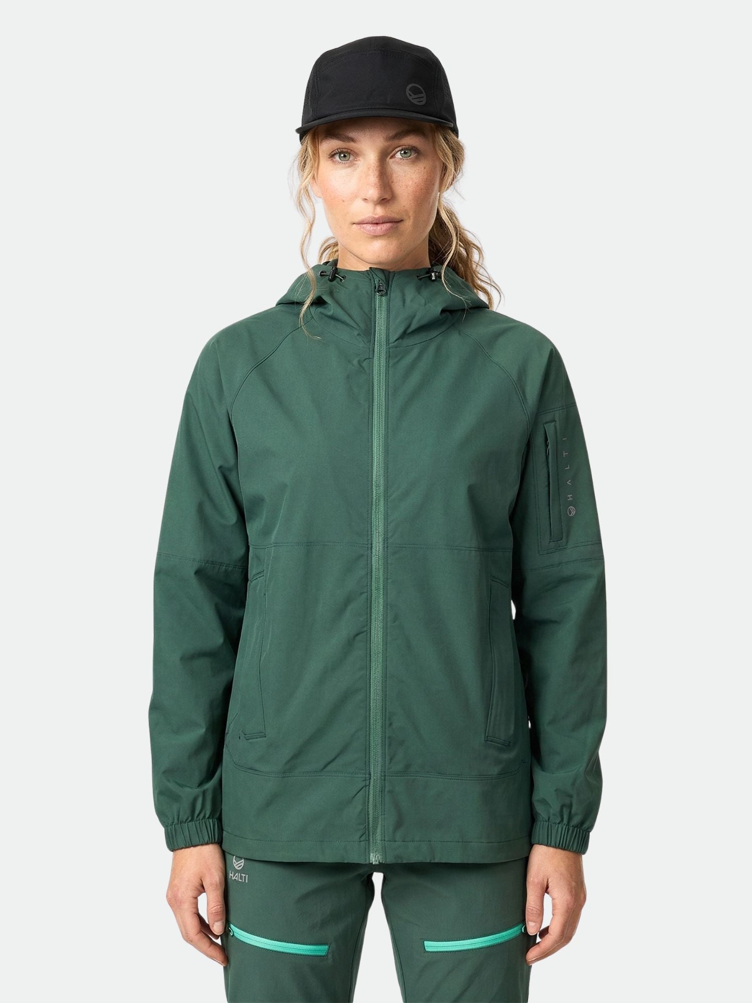 Naisella on yllään Halti Pallas Naisten Jogger Lite Takki, jossa on yhteensopivat housut ja musta lippis valkoista taustaa vasten.