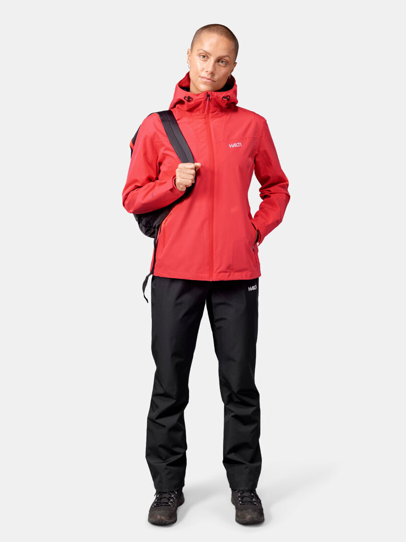 Henkilöllä on yllään punainen Halti Forter DX Kuoritakki Naisten, mustat housut, saappaat ja musta reppu tavallista taustaa vasten.