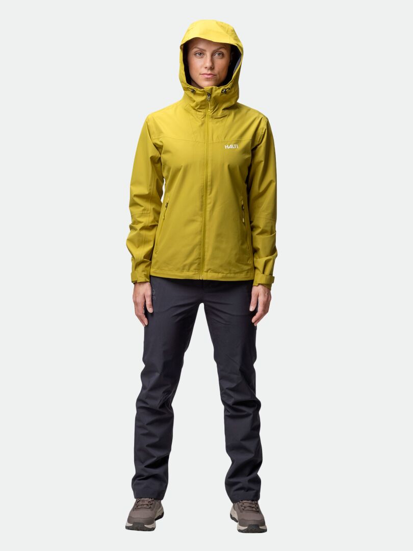 Henkilö pukeutuu Halti Forter DX Kuoritakki Naisten keltaiseen, tummiin housuihin ja ruskeisiin kenkiin, kasvot menosuuntaan.