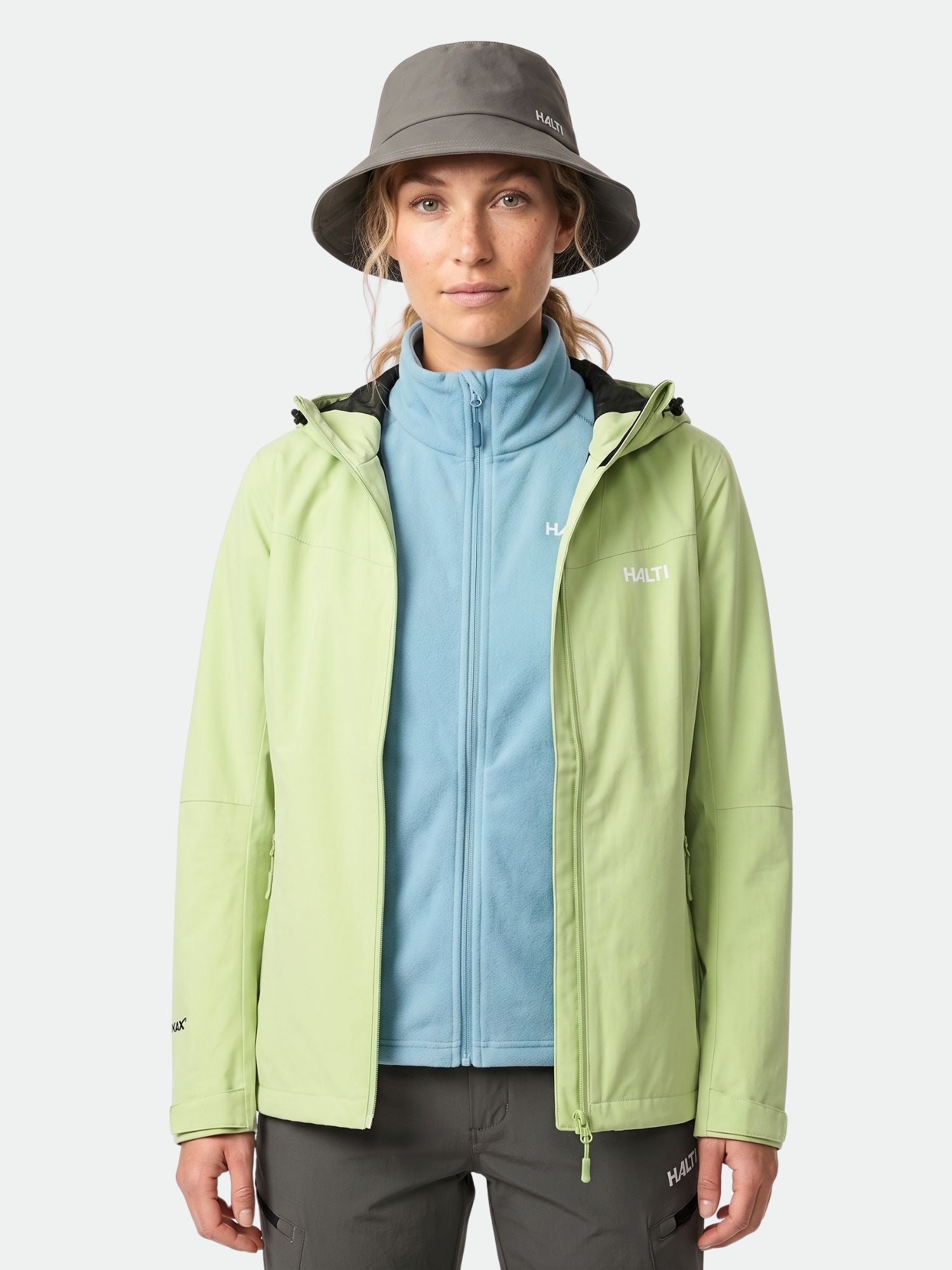 Nainen, jolla on harmaa kauhahattu, sininen fleece ja Halti Forter DX Kuoritakki Naisten seisoo kasvot menosuuntaan.