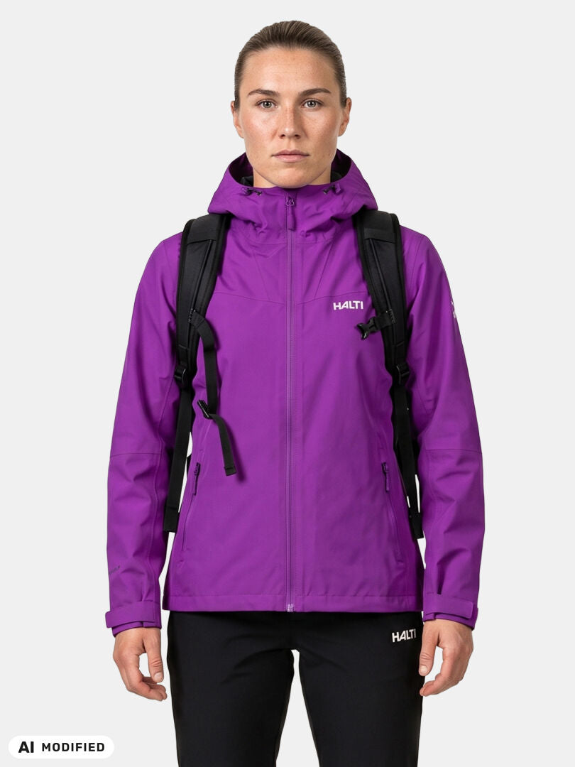 Nainen, jolla on yllään violetti Halti Forter DX Kuoritakki -takki ja mustat housut sekä musta reppu, valkoisella taustalla.