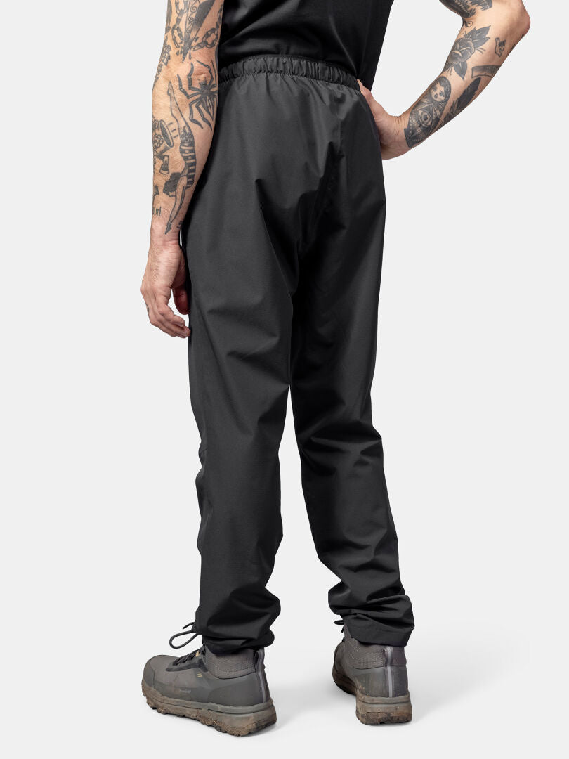 Henkilöllä on yllään Halti Forter DX Kuorihousut Miesten, harmaat vaelluskengät ja musta paita, kuvattu takaa.