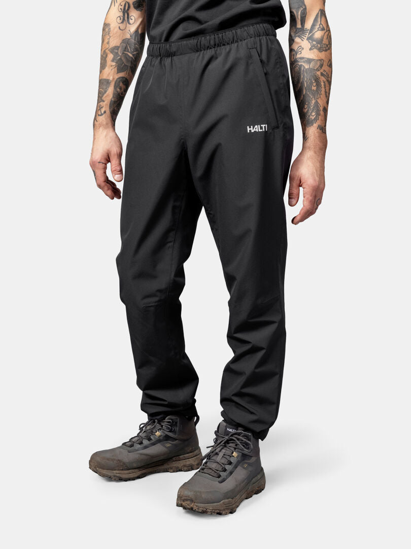 Henkilöllä on yllään musta Halti Forter DX Kuorihousut Miesten ja tummat vaelluskengät, seisoo vaaleaa taustaa vasten.