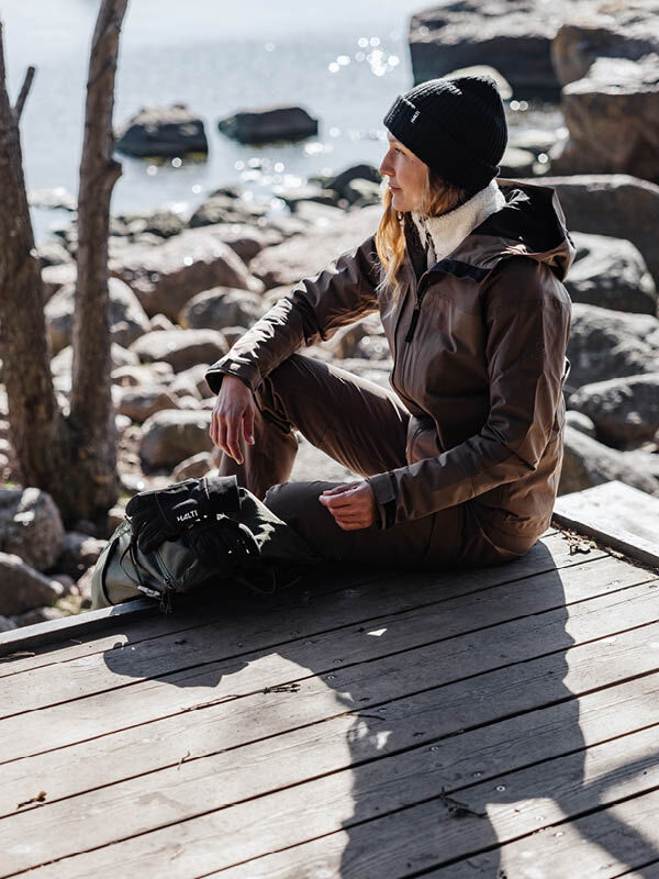 Nainen, jolla on yllään ruskea Halti Hiker II DrymaxX Kuoritakki Naisten -takki ja musta pipo, istuu puulaiturilla reppunsa vieressä.