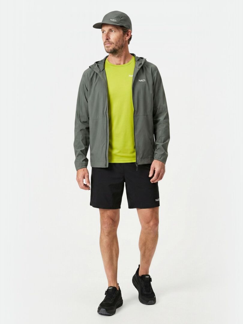 Halti Pisarat 2,5L DX Kuoritakki -mies seisoo mustissa shortseissa, mustissa lenkkareissa, lime-paidassa ja lippiksessä.
