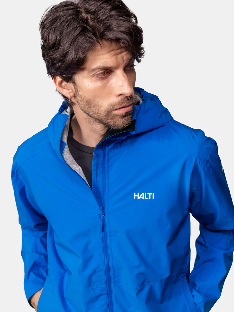 Miehellä on päällään sininen Halti Pisarat 2,5L DX Kuoritakki Miesten huppu, katselee sivulle tavallista taustaa vasten.