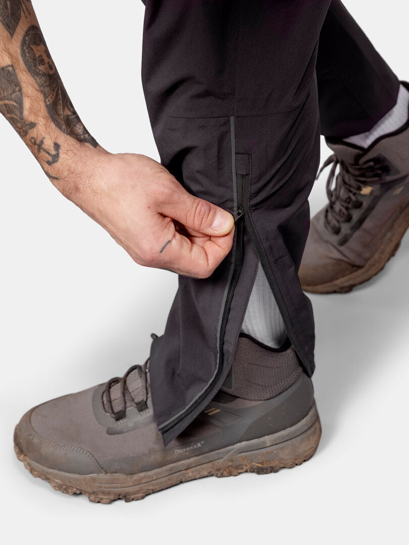 Henkilö avaa Halti Pisarat 2,5L DX Kuorihousut Miesten ruskeiden vaelluskenkien päällä, näyttää tatuoidun kyynärvarren ja valkoiset sukat.