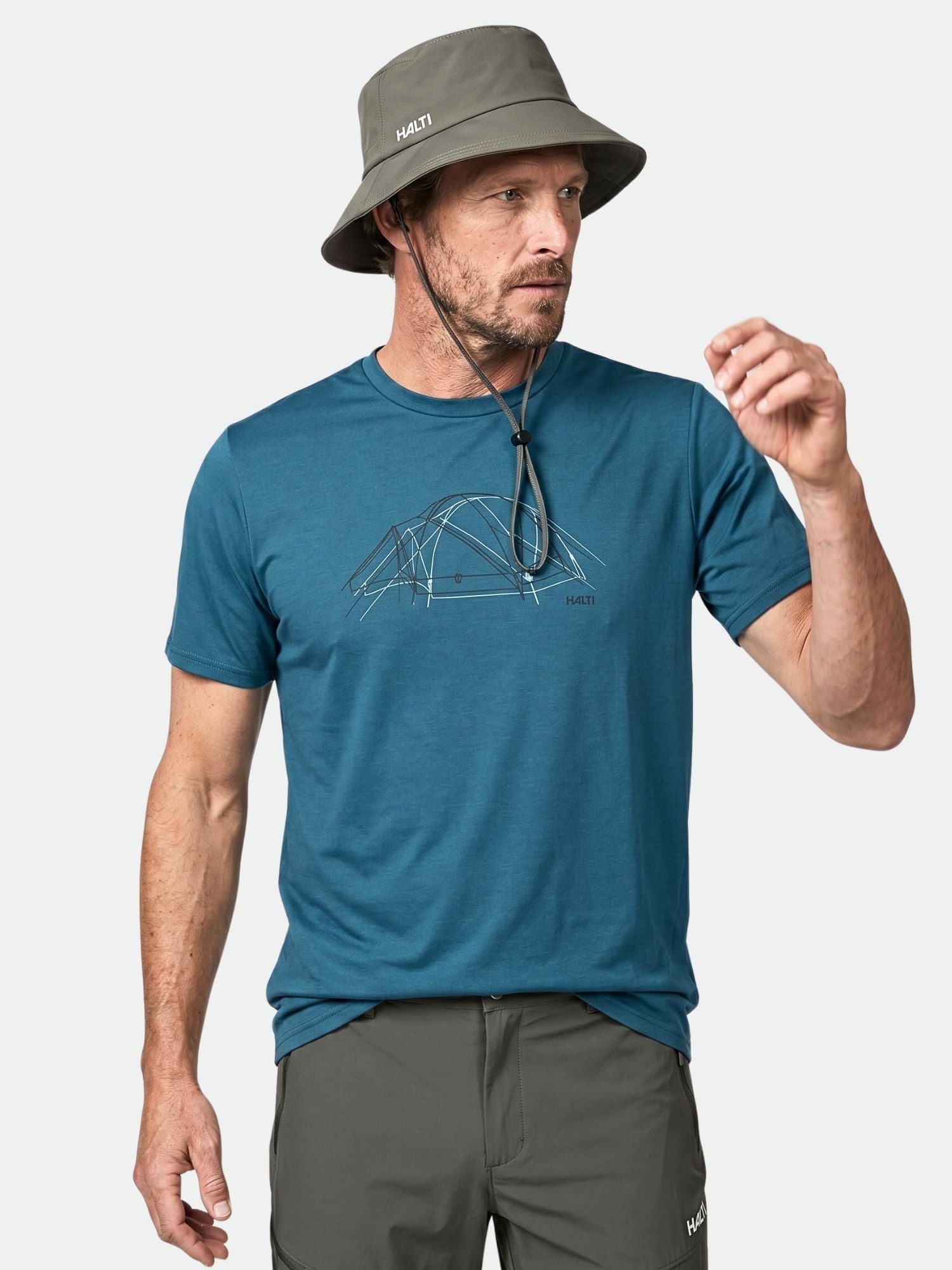 Mies, jolla on harmaa hattu ja Halti Kalteva T-paita Miesten teltta-grafiikalla, seisoo valkoista taustaa vasten.