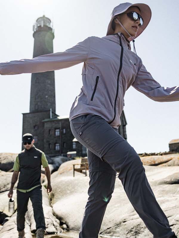 Kaksi ihmistä Halti Pallas Cool Stretch Zip-off Housut Naisten vaatteissa kävelee kallioilla korkean majakan luona kirkkaalla taivaalla.