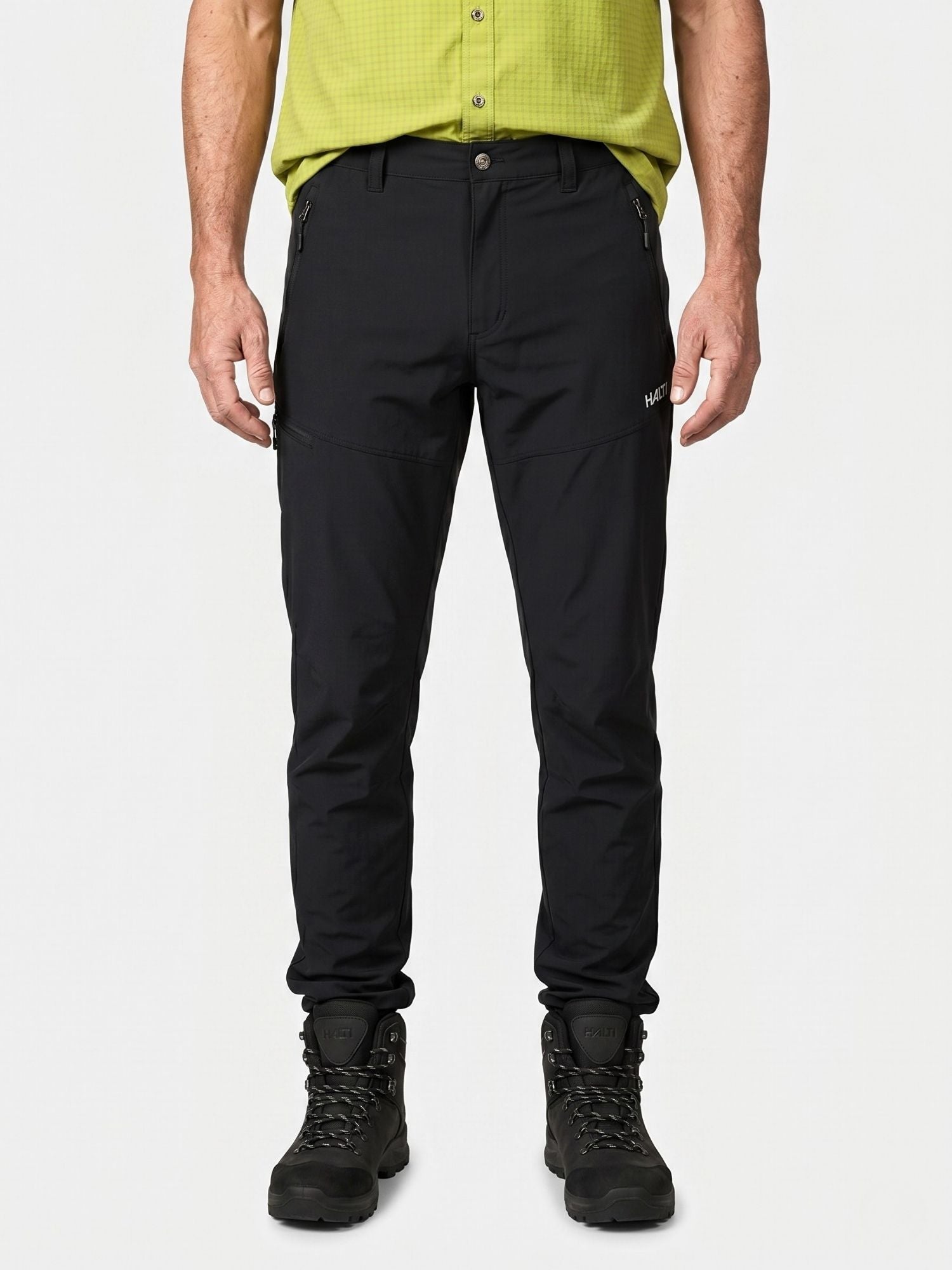 Henkilö, jolla on mustat Halti Pallas Cool X-stretch Housut Miesten, mustat saappaat ja vaaleanvihreä paita, seisoo valkoista vasten.