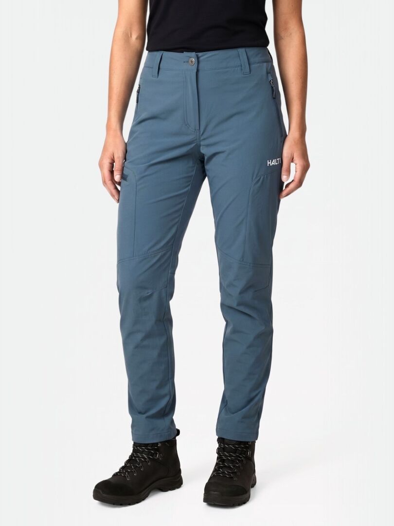 Henkilö, jolla on siniset Halti Pallas Cool X-stretch Housut Naisten, mustat saappaat ja musta paita, seisoo valkoista taustaa vasten.