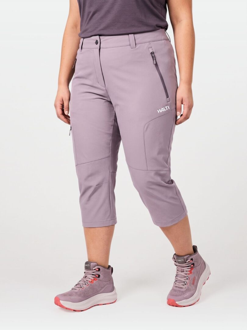 Henkilöllä on yllään Halti Pallas Cool X-stretch Caprit Naisten Plus ja harmaat vaelluskengät, joissa on vaaleanpunainen pohja, yksiväristä taustaa vasten.