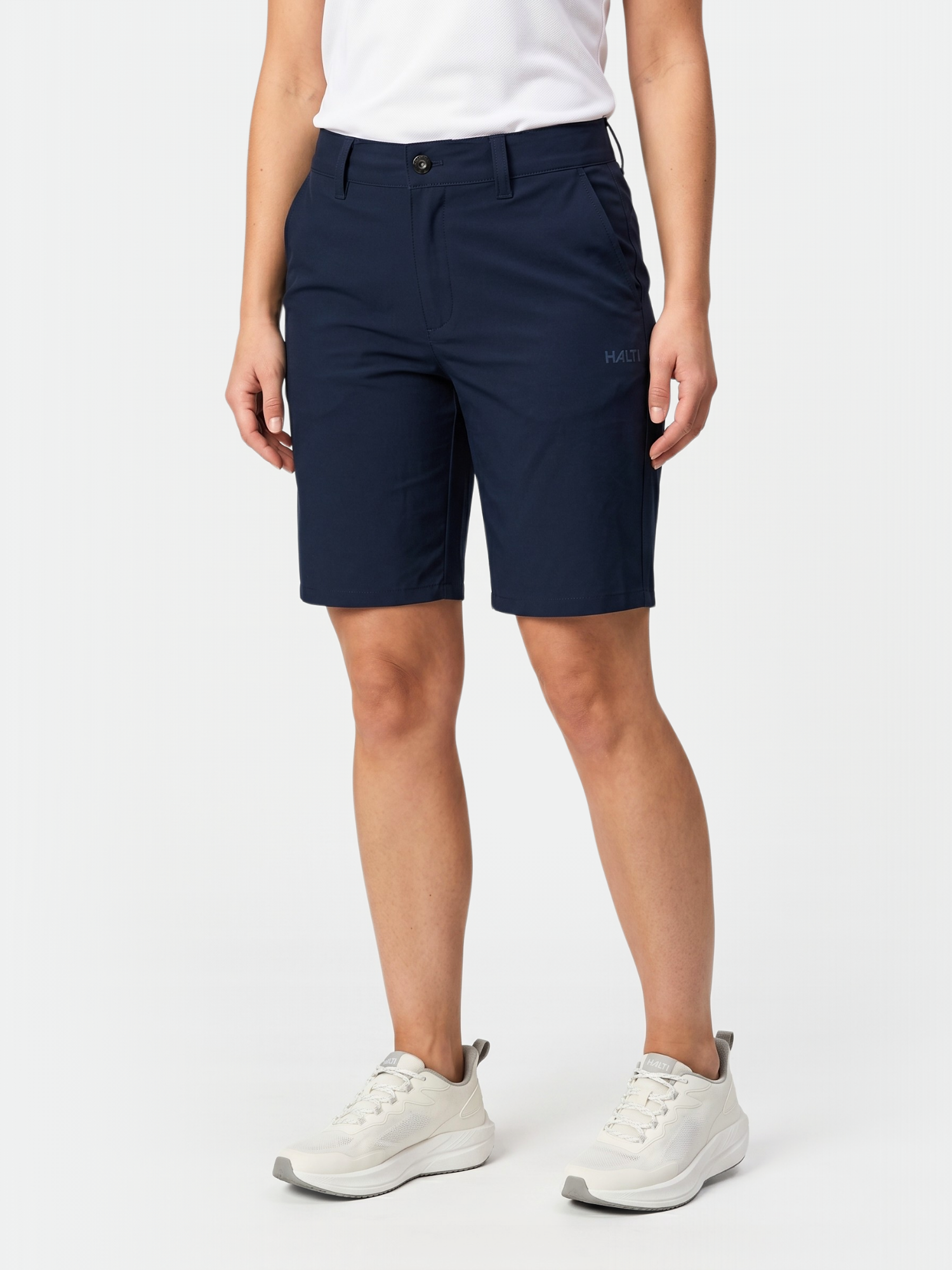 Henkilö, jolla on Halti Drive X-stretch Shortsit Naisten, valkoinen paita ja lenkkarit, seisoo valkoista taustaa vasten.