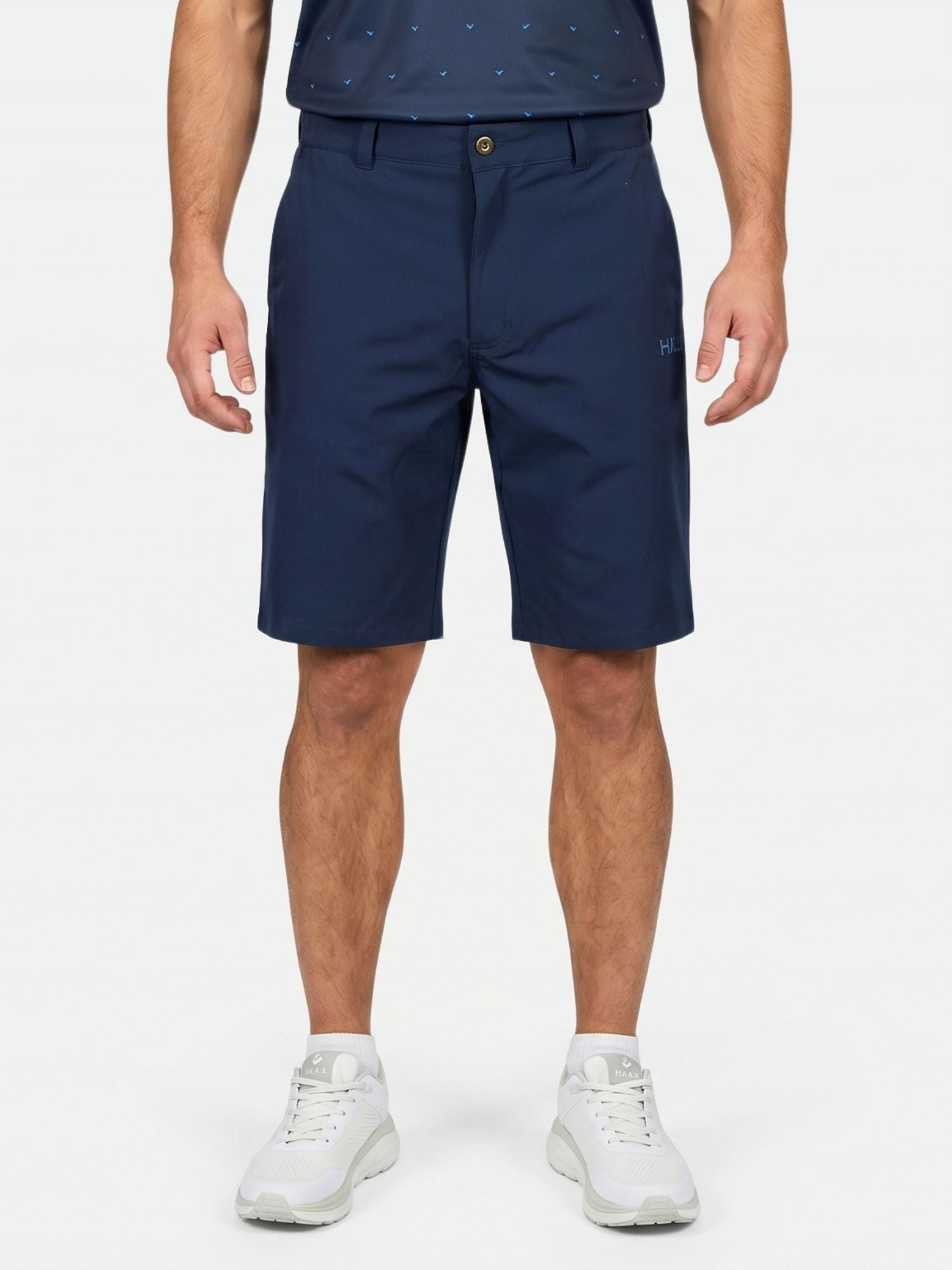 Henkilö on pukeutunut Halti Drive X-stretch Shortsit Miesten -shortseihin, tummaan paitaan ja valkoisiin lenkkareihin vaaleaa, yksiväristä taustaa vasten.