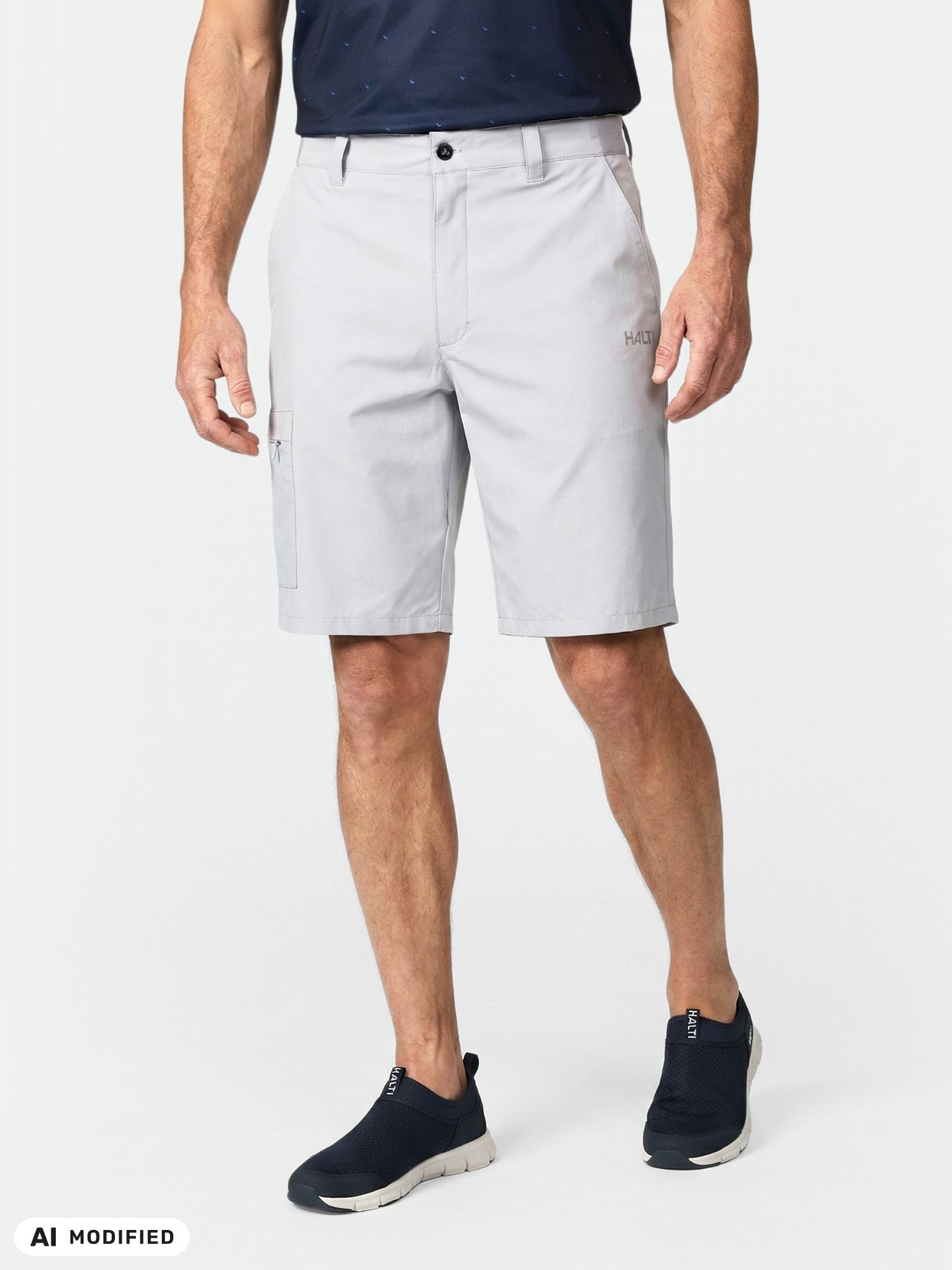 Mies, jolla on yllään Halti Drive X-stretch Shortsit Miesten -shortsit, tummansininen paita ja mustat slip-on-kengät, seisoo yksivärisen taustan edessä.
